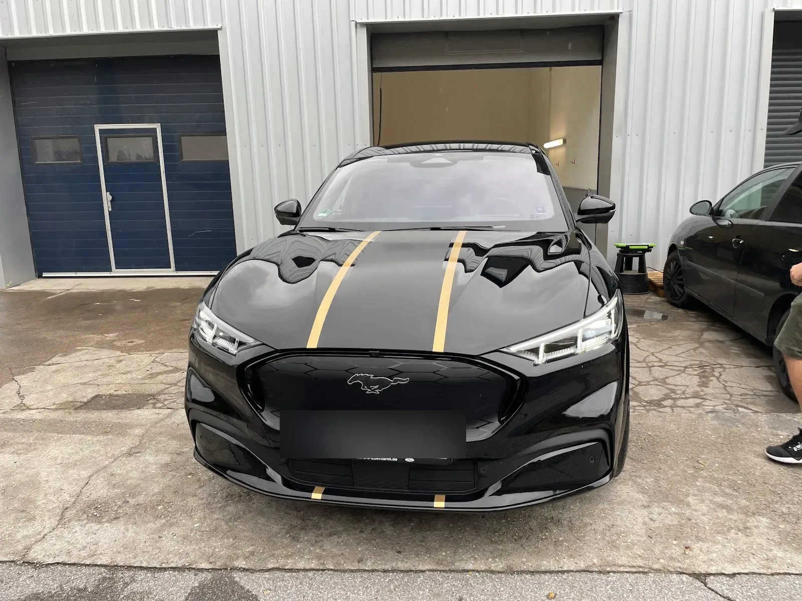 Schwarzer Ford Mustang Mach-E mit goldenen Streifen frontal vor der Halle, Lack frisch aufbereitet und stark spiegelnd.