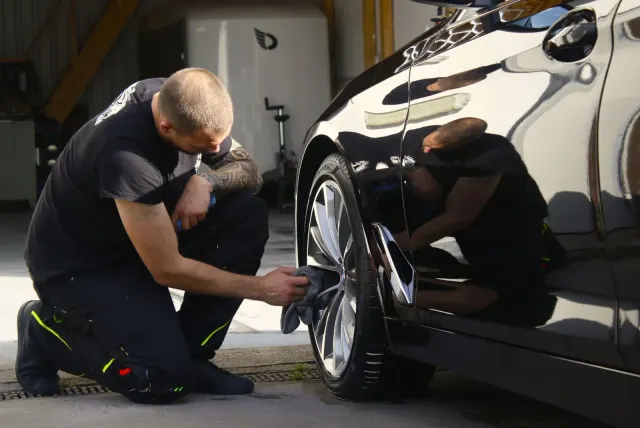 Mitarbeiter kniet an einem schwarzen BMW 5er und poliert die Felge mit einem Mikrofasertuch, Lack spiegelt die Szene.