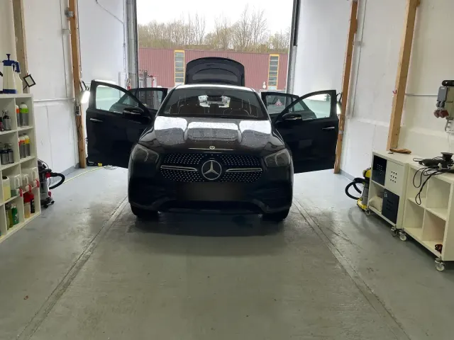 Schwarzer Mercedes GLE Coupe mit geoeffneten Tueren und Heckklappe in der hellen Aufbereitungshalle, Pflegemittel-Regal und Sauger im Bild.