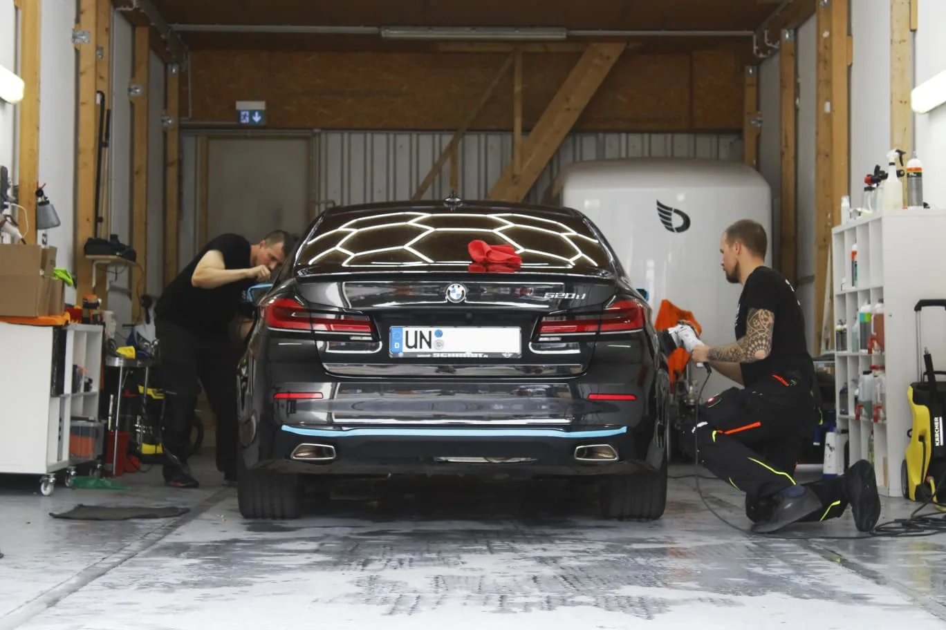 Zwei Mitarbeiter polieren einen schwarzen BMW 520d in der Aufbereitungshalle unter Hexagon-Licht, Heckansicht mit nasser Bodenflaeche.
